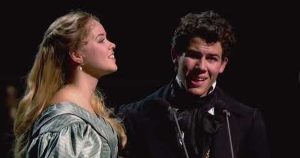Two performers in period-style costumes sing together on a dark stage, captured in a dramatic live musical theater moment.