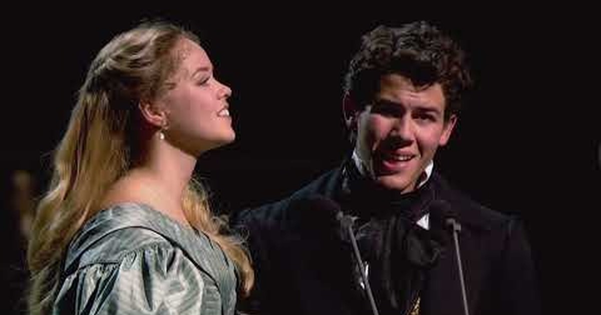 Two performers in period-style costumes sing together on a dark stage, captured in a dramatic live musical theater moment.